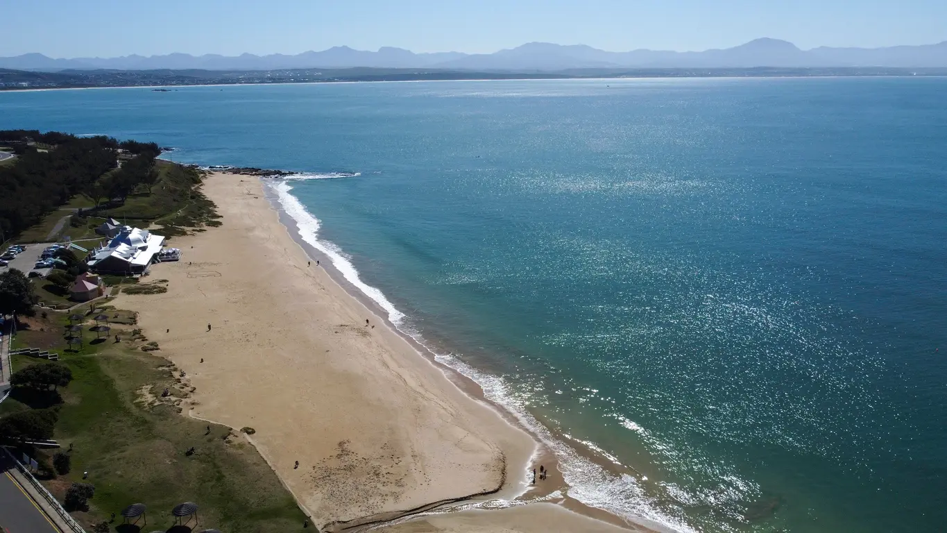Aerial view of Santos Beach in Mossel Bay with calm turquoise water, a wide sandy shoreline and the Outeniqua Mountains in the distance.