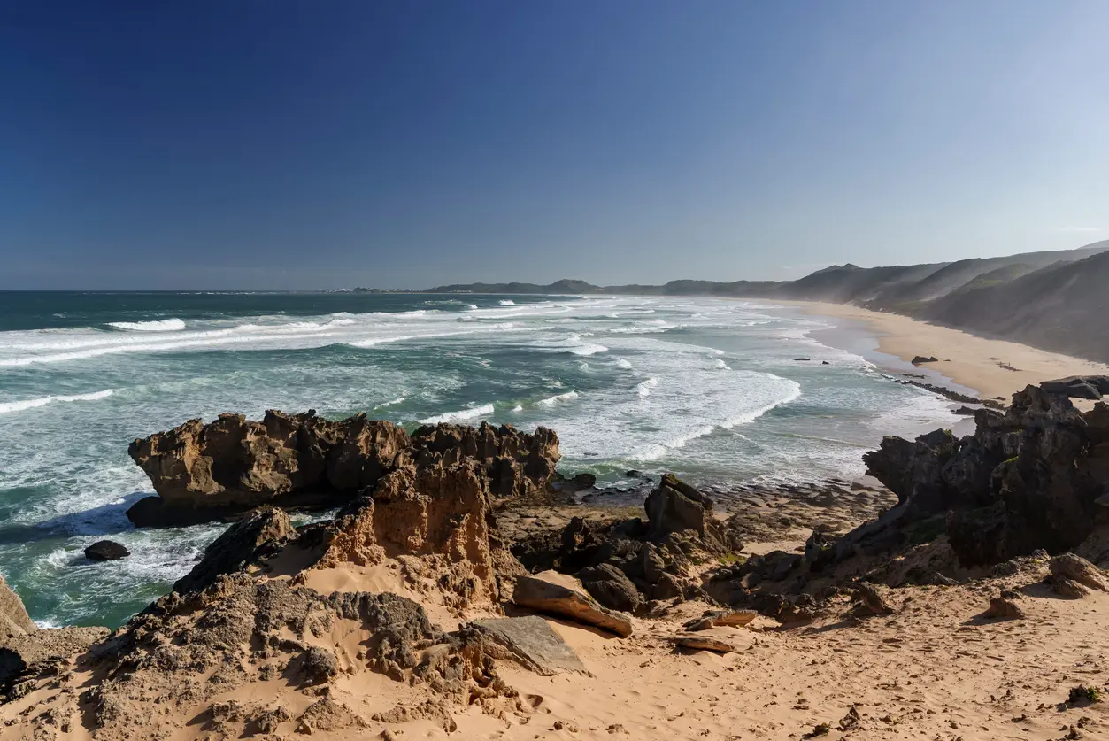 Elevated view of Brenton Beach showing rocky coastal cliffs, breaking ocean waves and a long sandy shoreline stretching along the Garden Route near Knysna, South Africa.