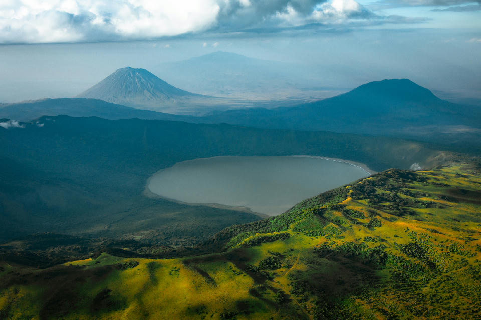 Aerial photograph of Empakaai Crater in Tanzania, showing the forested volcanic caldera and crater lake with Ol Doinyo Lengai rising beyond the Great Rift Valley landscape.