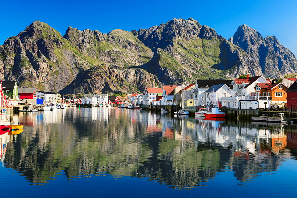 Colourful fishing houses and boats reflected in the harbour at Henningsvær on Austvågøya in the Lofoten Islands, with steep granite peaks rising behind in Nordland, Norway.
