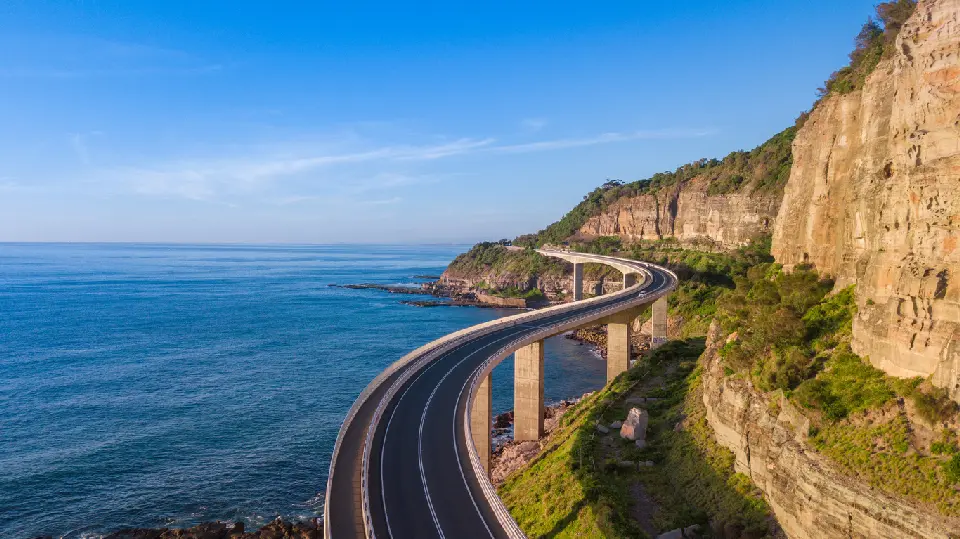 Sweeping aerial view of Sea Cliff Bridge curving between steep sandstone cliffs and the blue Pacific Ocean on the Illawarra coast.