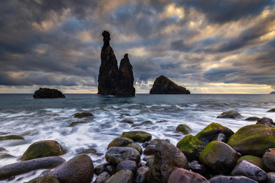Ilhéu da Ribeira da Janela rock stacks silhouetted against dramatic sunset clouds, waves washing over mossy volcanic stones on the Madeira coastline, Portugal.
