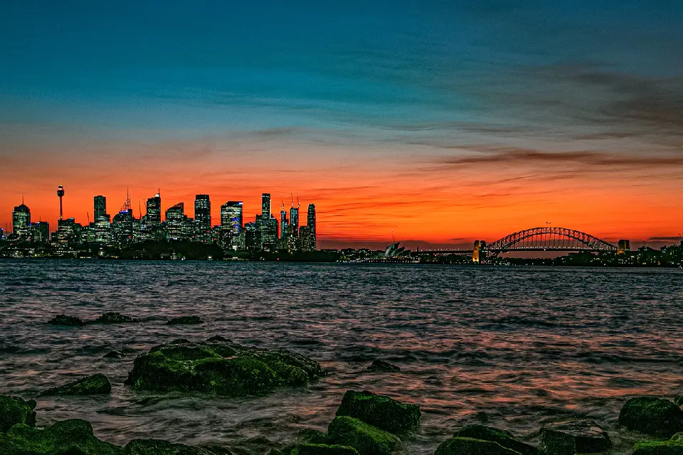 Fiery orange sunset over Sydney Harbour from Bradleys Head, with the CBD skyline, Opera House and Harbour Bridge beyond the rocky shoreline.