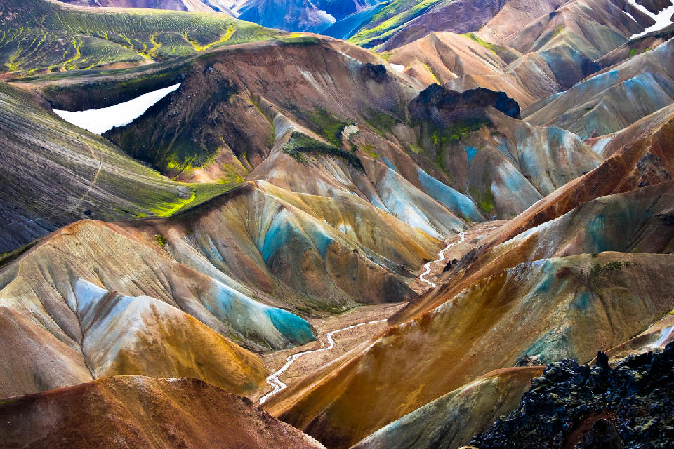 Multicolored rhyolite hills and a narrow river in the Iceland Highlands, showing layered volcanic slopes with patches of green moss and lingering snow.