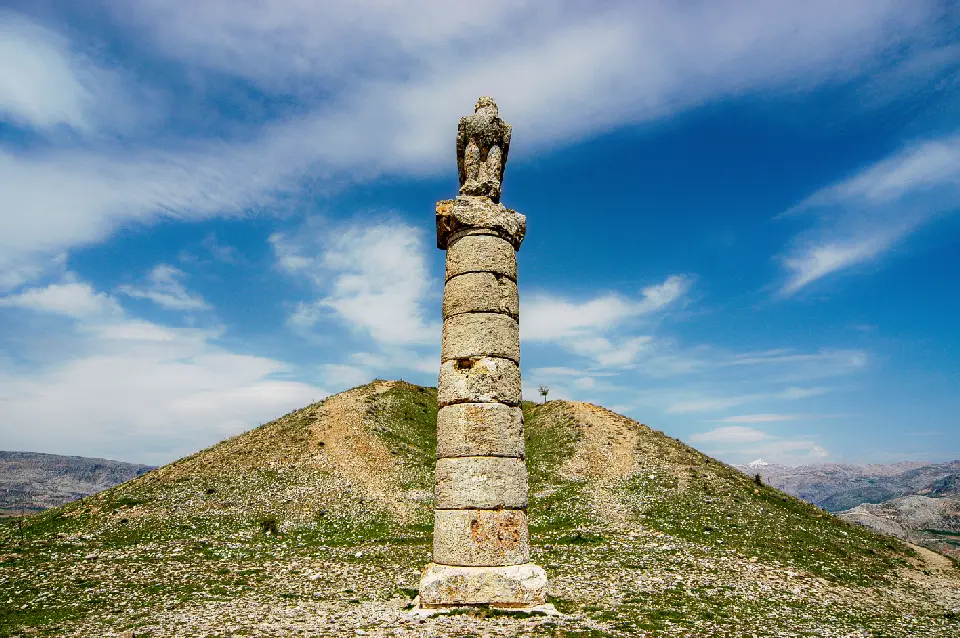 Ancient stone column topped by an eagle statue beside the Karakuş Tumulus in the mountainous landscape of Adıyaman, Turkey.