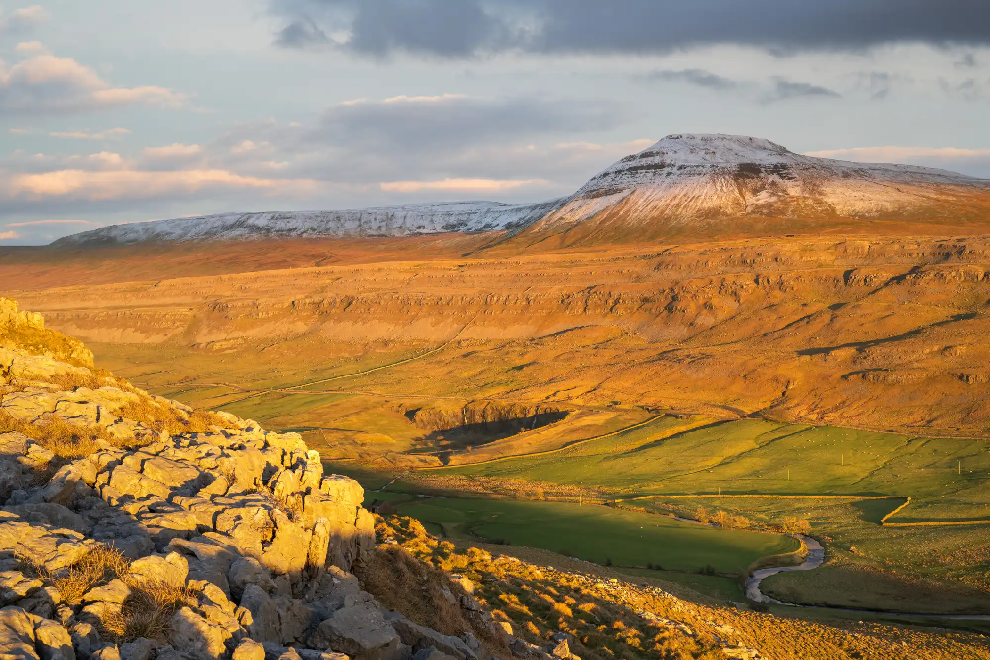 Snow-capped Ingleborough glowing in golden evening sunlight above limestone rocks and a winding river valley in the Yorkshire Dales, England.