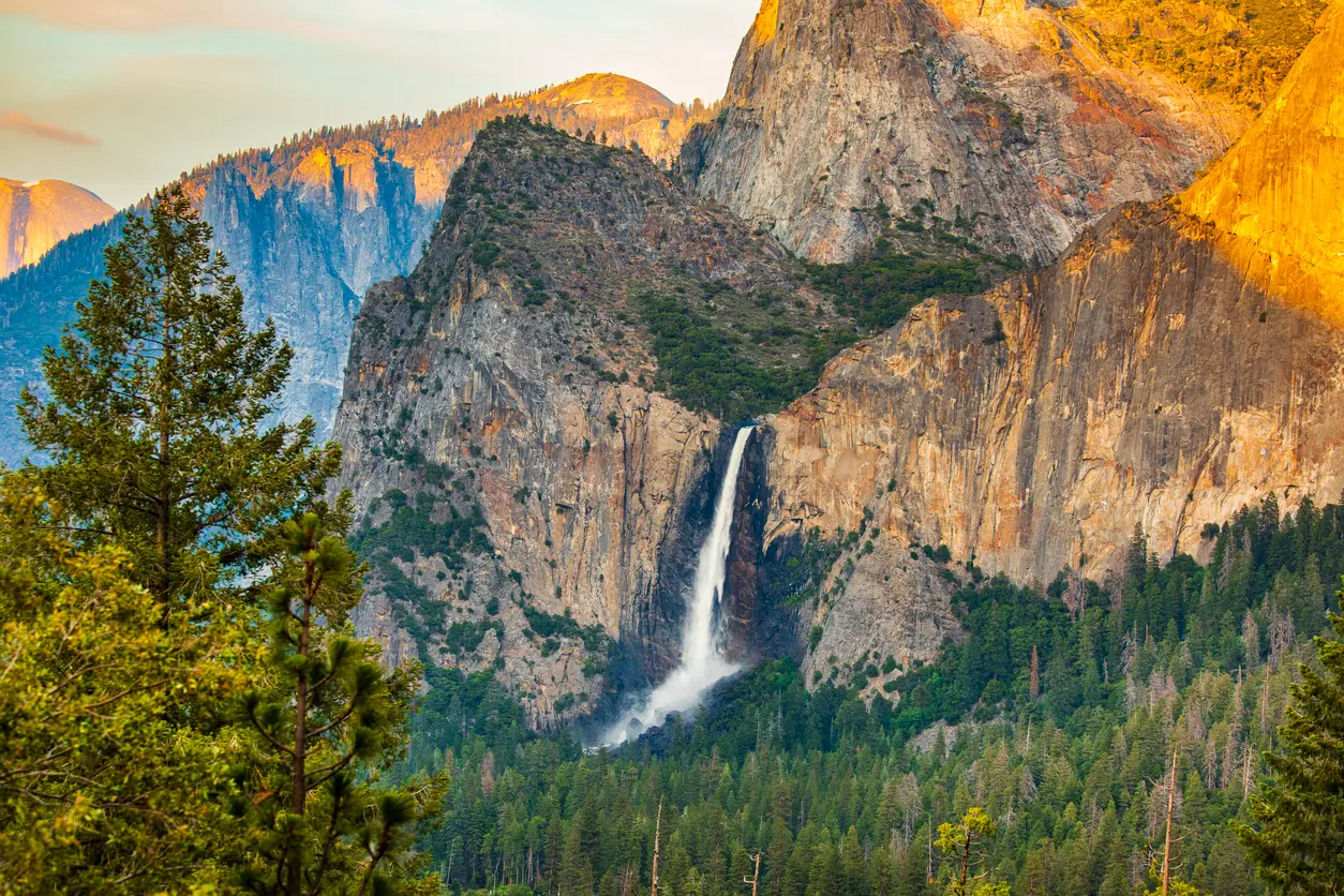 Bridalveil Fall Yosemite with tall waterfall dropping from granite cliffs above dense pine forest in warm afternoon light.