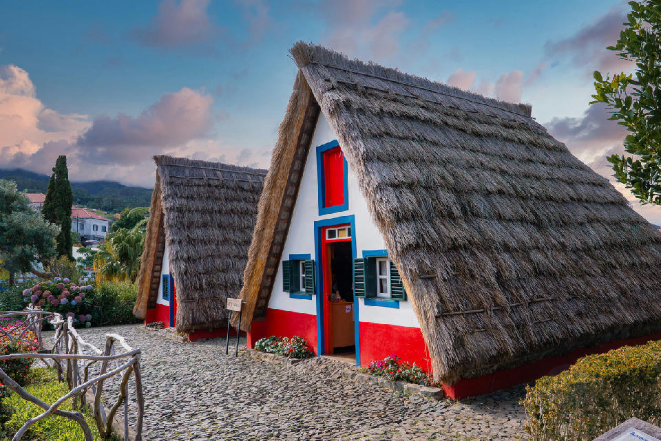 Traditional A-frame thatched houses in Santana, Madeira, with white walls and red bases beside a cobblestone path under a pastel evening sky.