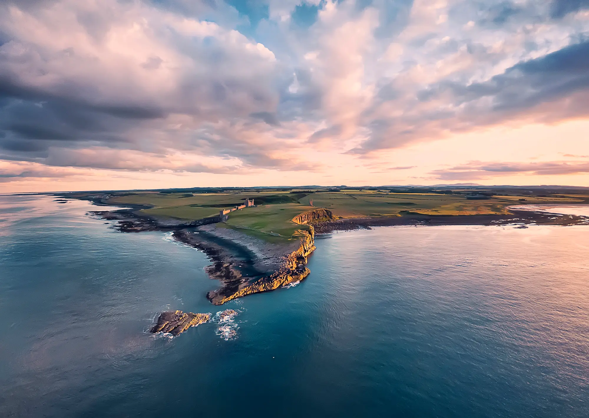 Aerial view of Dunstanburgh Castle on a rocky Northumberland headland, surrounded by the North Sea and green fields beneath dramatic pink and purple sunset clouds.