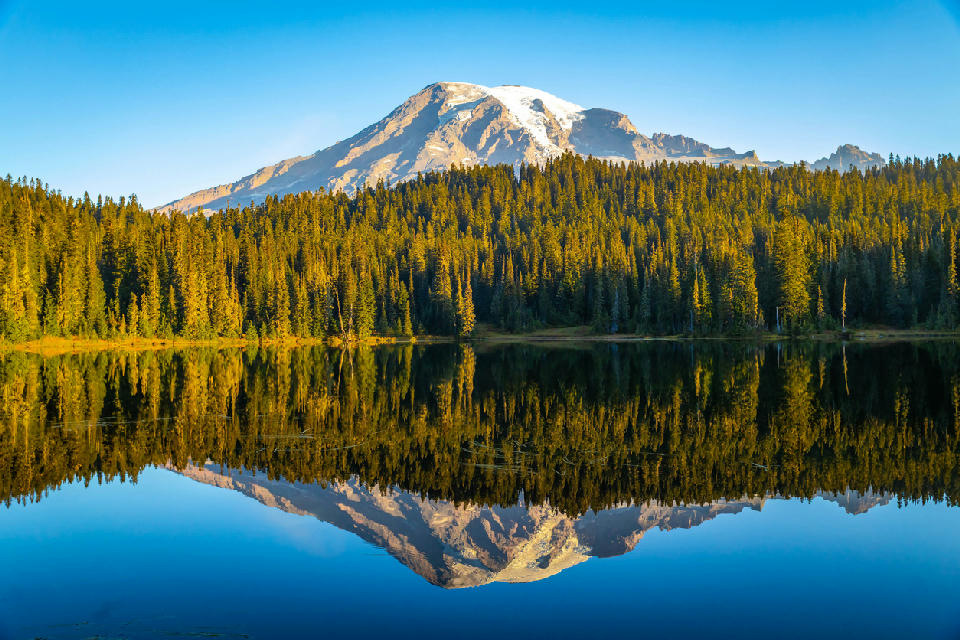 Mount Rainier reflected in Reflection Lakes with calm water, forest shoreline, and snow-covered peak in Mount Rainier National Park.