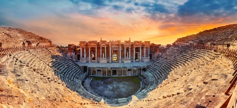 Panoramic view of the ancient Roman Theatre of Hierapolis, with its tiered stone seating and monumental stage façade beneath a dramatic sunset sky.