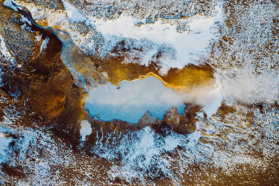 Top-down drone view of a steaming geothermal pool surrounded by yellow sulphur deposits and snow at the Seltún geothermal area in Krýsuvík, Iceland.