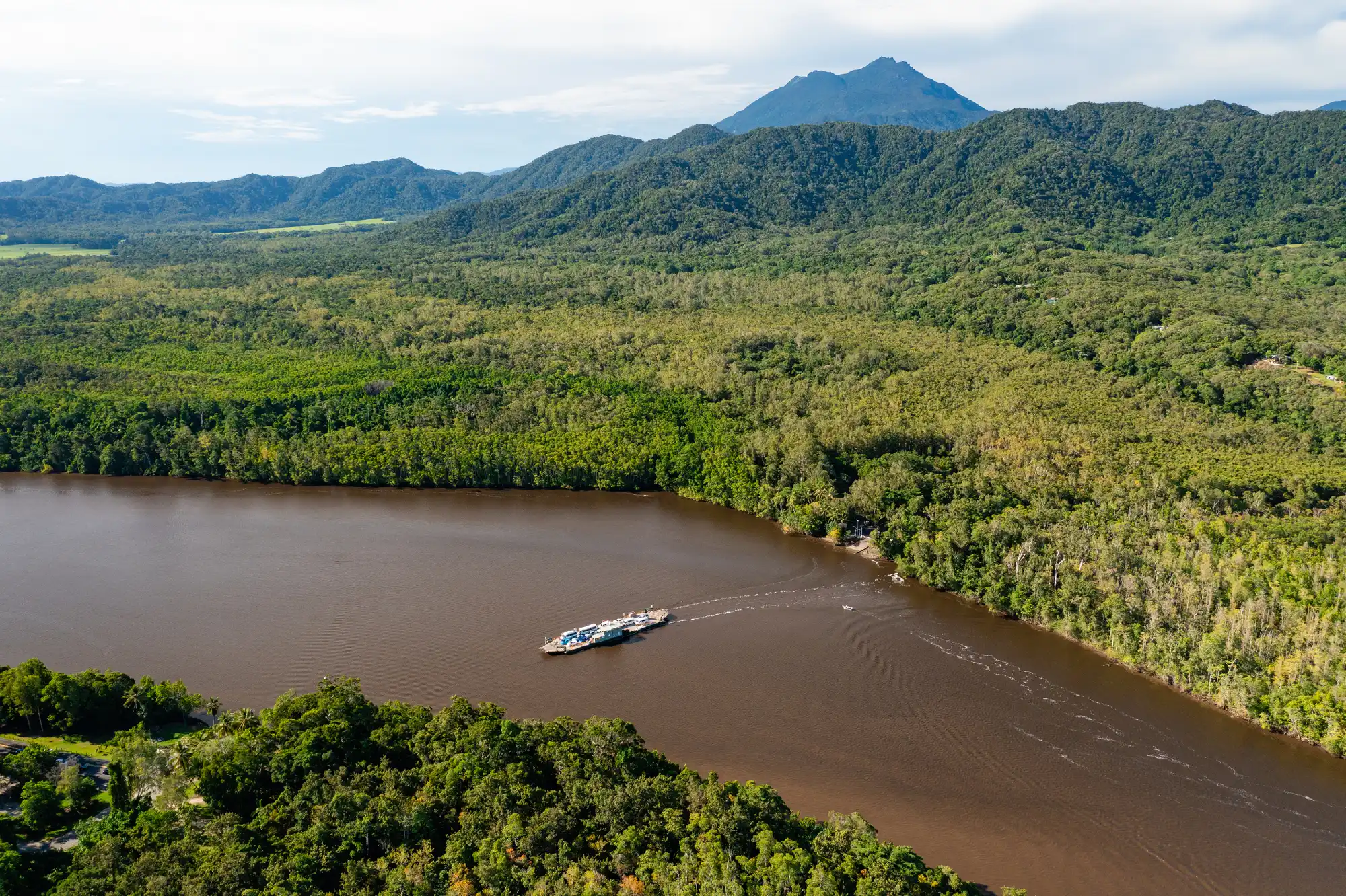 A vehicle ferry creates small ripples as it crosses a wide brown river running through a dense, vibrant green jungle.