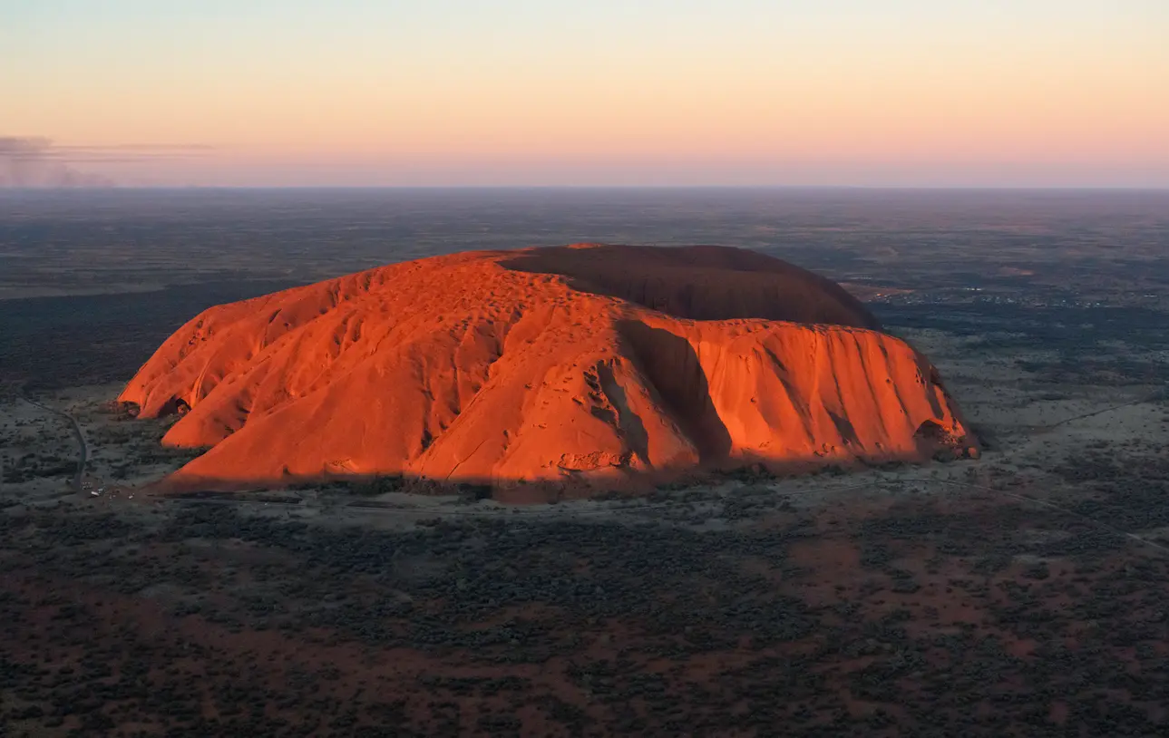 Aerial view of Uluru glowing red in evening light above the surrounding desert in Uluru-Kata Tjuta National Park, Australia.