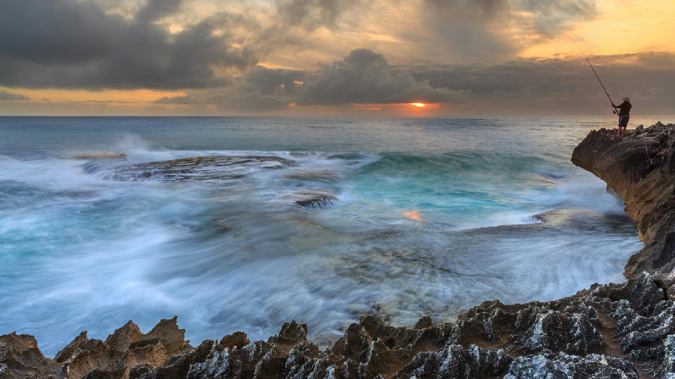 Fisherman on rocky cliffs above turbulent ocean waves near Arniston on the Western Cape coast of South Africa at sunrise.