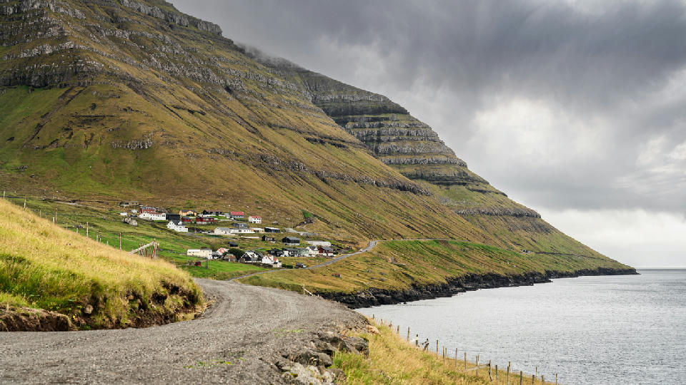 Empty gravel road along the fjord approaching Kunoy village at the base of tall terraced basalt mountains on Kunoy Island, Faroe Islands.