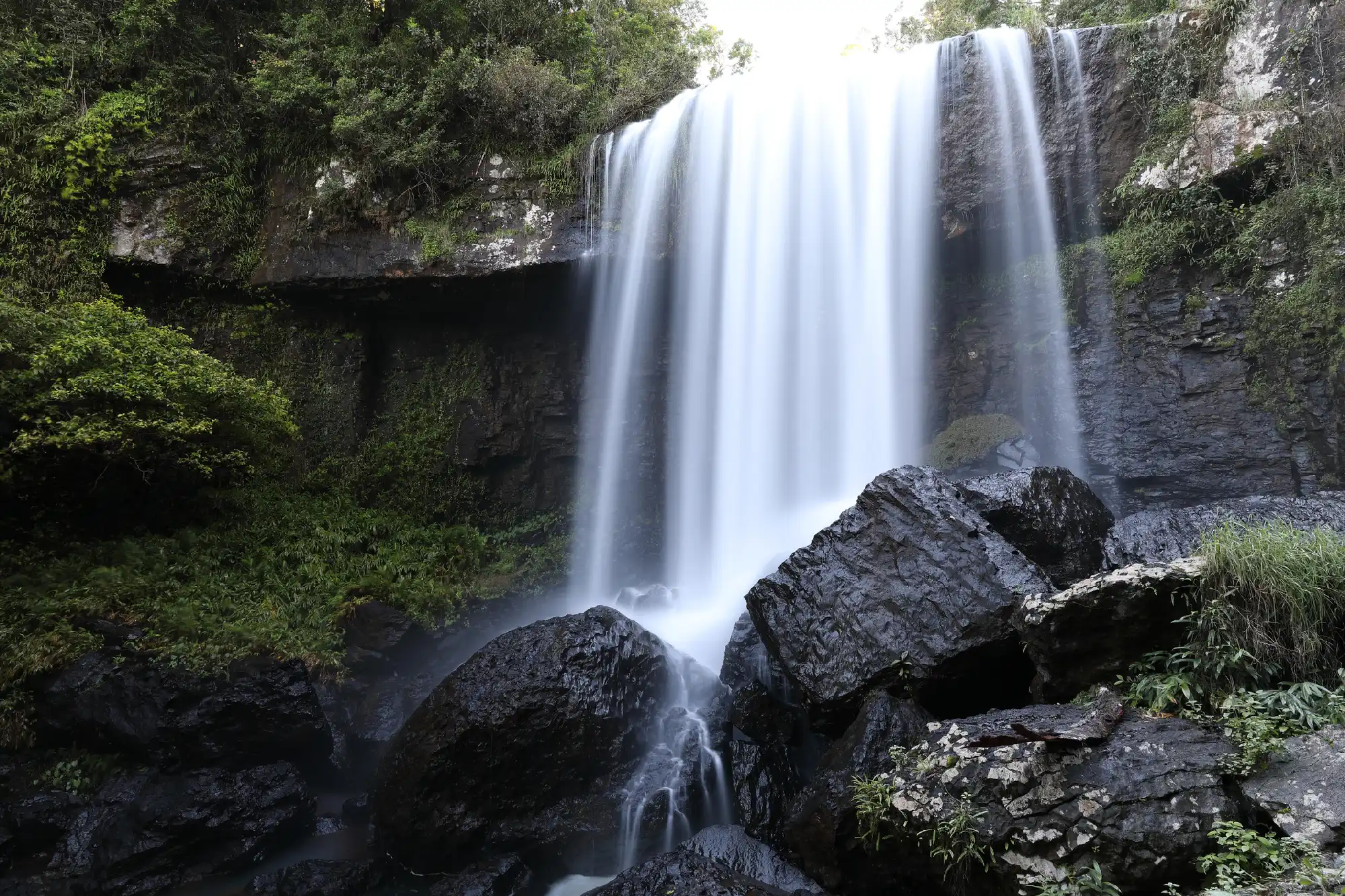 Frontal view of Zillie Falls cascading over a broad rocky cliff into a gorge filled with large dark boulders and lush rainforest in Queensland, Australia.