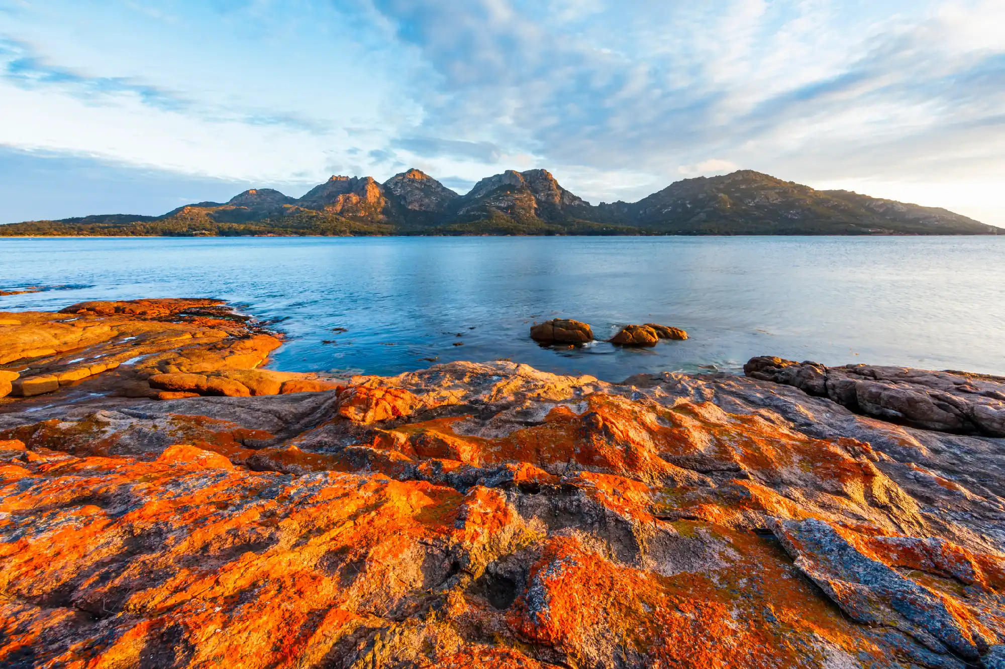 Orange-lichen-covered granite rocks beside calm blue water with The Hazards mountain range glowing in evening light beyond, Freycinet National Park, Tasmania.
