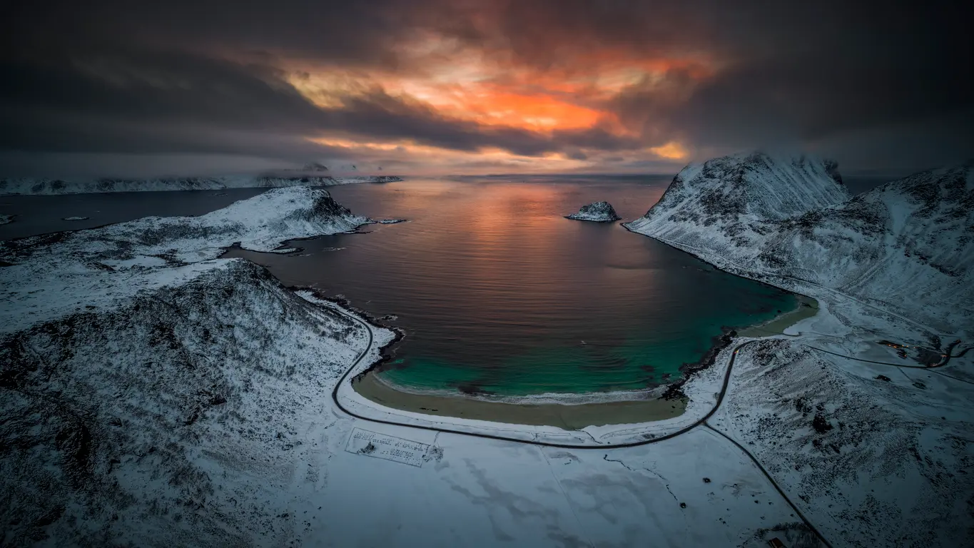 Drone view of Haukland and Vik Beach on Vestvågøya in winter, with snow-covered mountains surrounding a turquoise bay under a dramatic red-orange twilight sky in the Lofoten Islands, Norway.