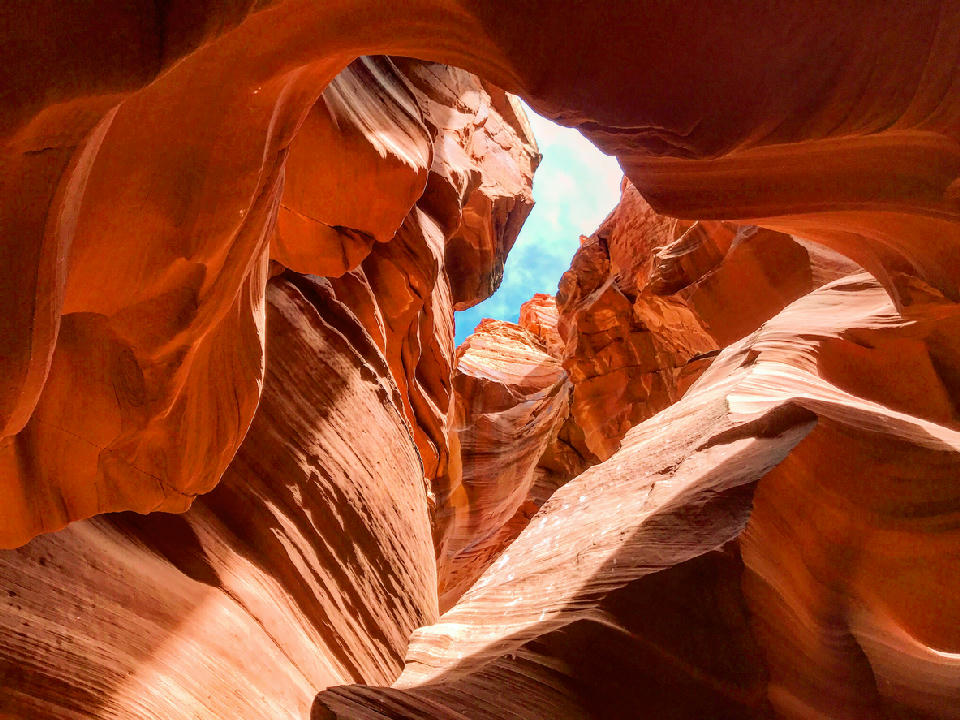 Canyon X in Page, Arizona showing upward view of narrow slot canyon with flowing sandstone walls and a small opening to blue sky above.