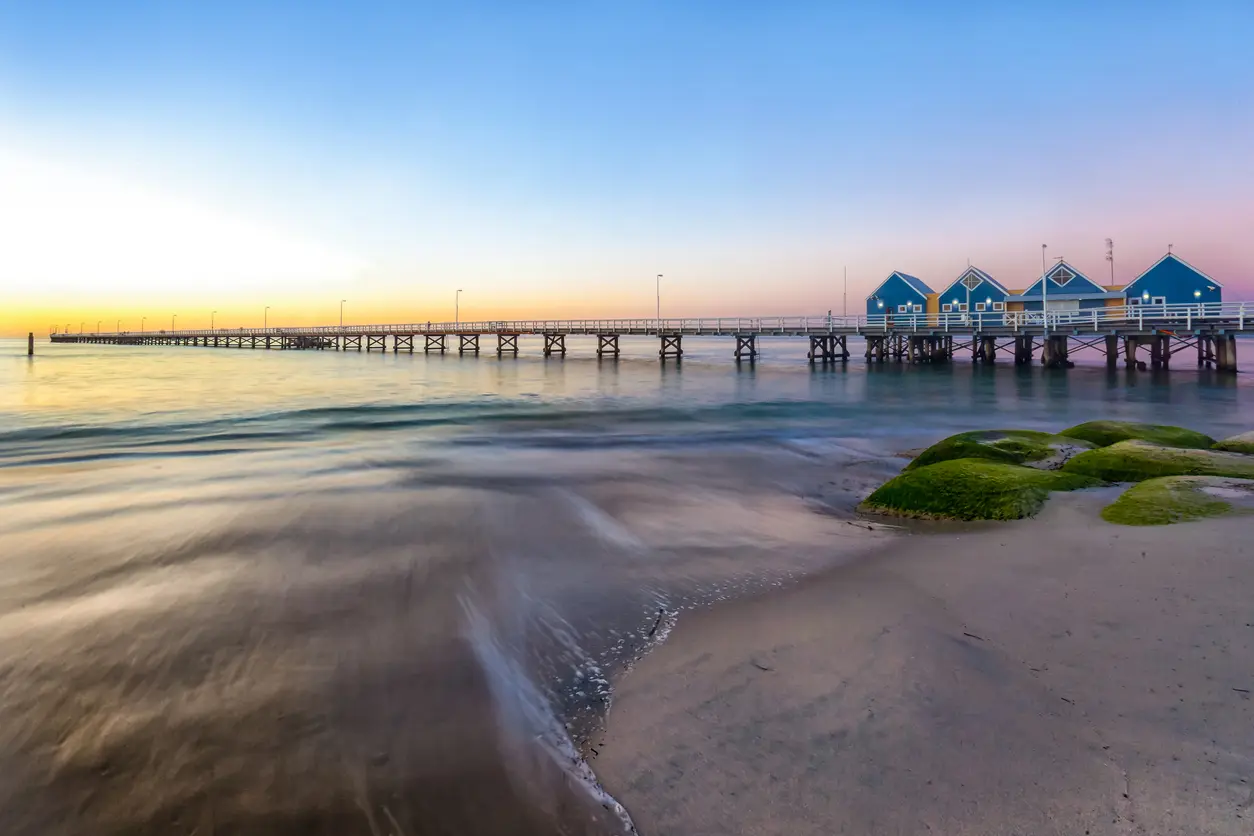 Busselton Jetty extending across calm water at sunset, with its blue buildings and a pastel sky over Geographe Bay.