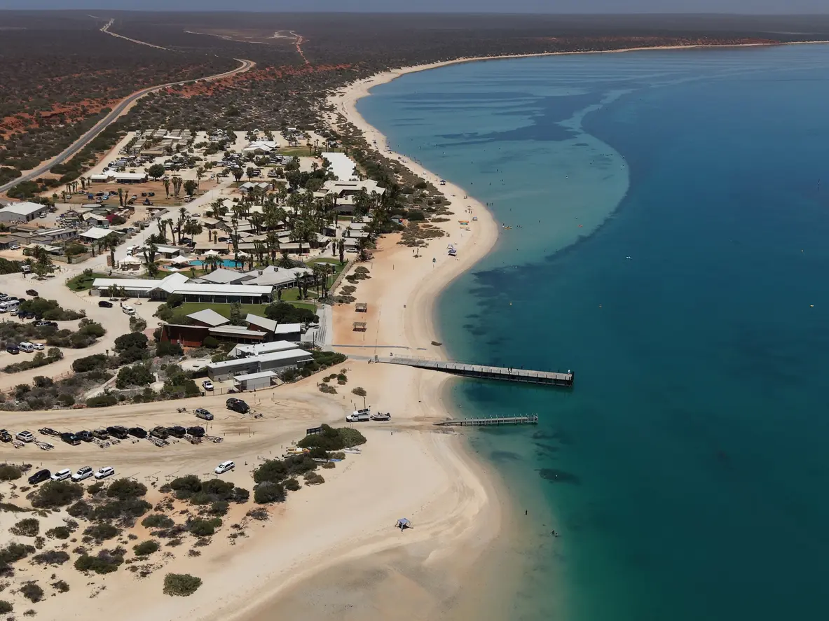 Aerial view of Monkey Mia showing the curved sandy beach, jetty, coastal buildings and turquoise waters of Shark Bay.