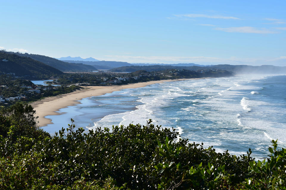 View over Lookout Beach in Plettenberg Bay showing a long sandy shoreline, breaking waves and distant mountains along the Garden Route coast.