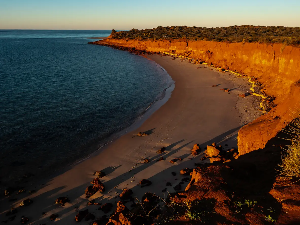 Warm sunset light illuminates red coastal cliffs above a curved sandy beach and deep blue water.