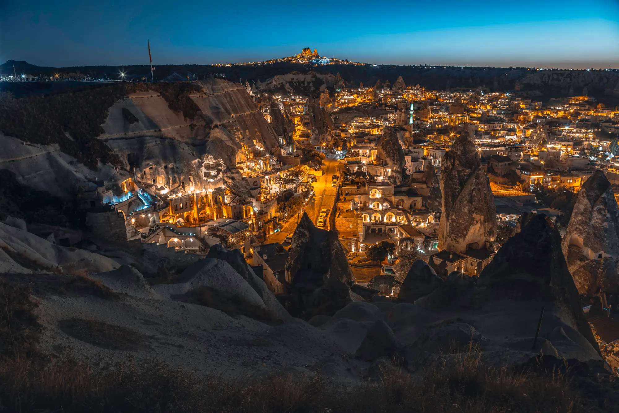 Night view of Göreme Town in Cappadocia featuring illuminated cave hotels, fairy chimneys and Uçhisar Castle on the horizon.