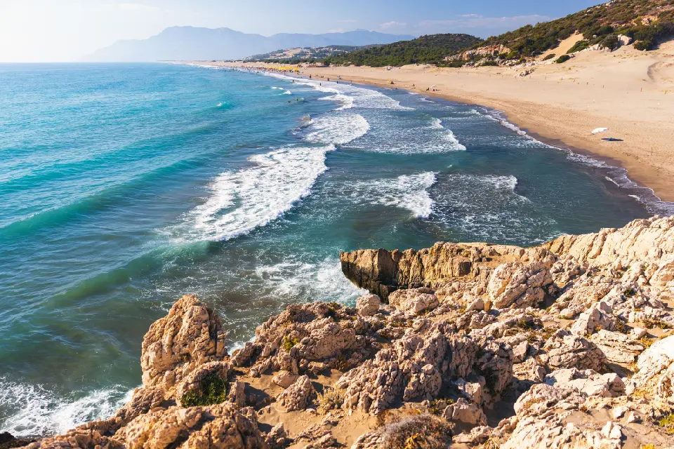 Elevated coastal view of Patara Beach, with turquoise waves rolling toward a long sandy shoreline, rugged limestone rocks in the foreground and hazy mountains in the distance.