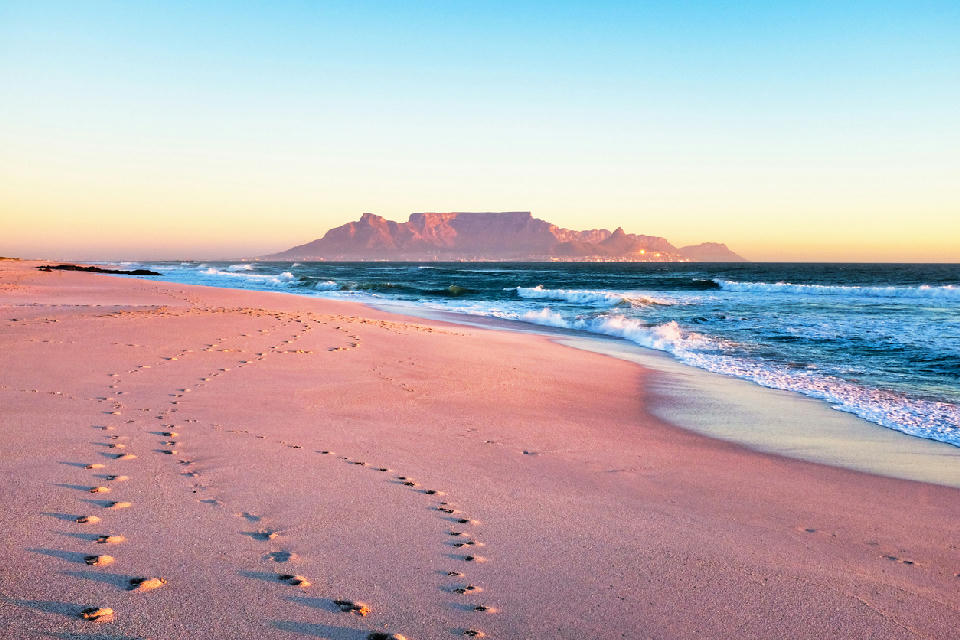 Table Mountain viewed from Bloubergstrand beach at sunset with waves rolling onto the shore and footprints in the sand, Cape Town, South Africa.