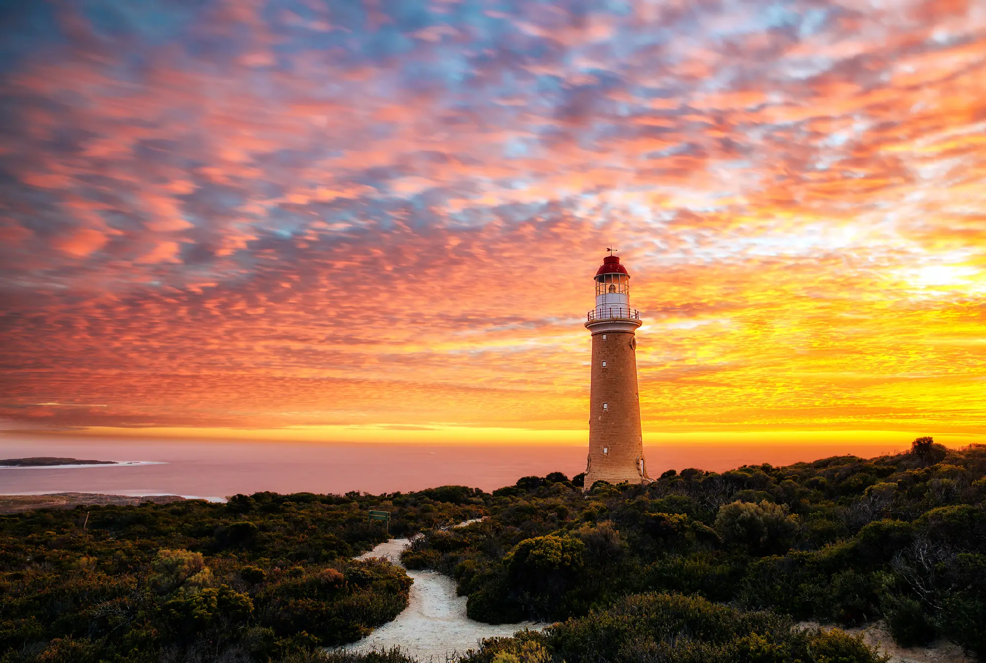Cape du Couedic Lighthouse at sunset on Kangaroo Island, with a winding sandy path, dark coastal vegetation and vivid orange-pink clouds.