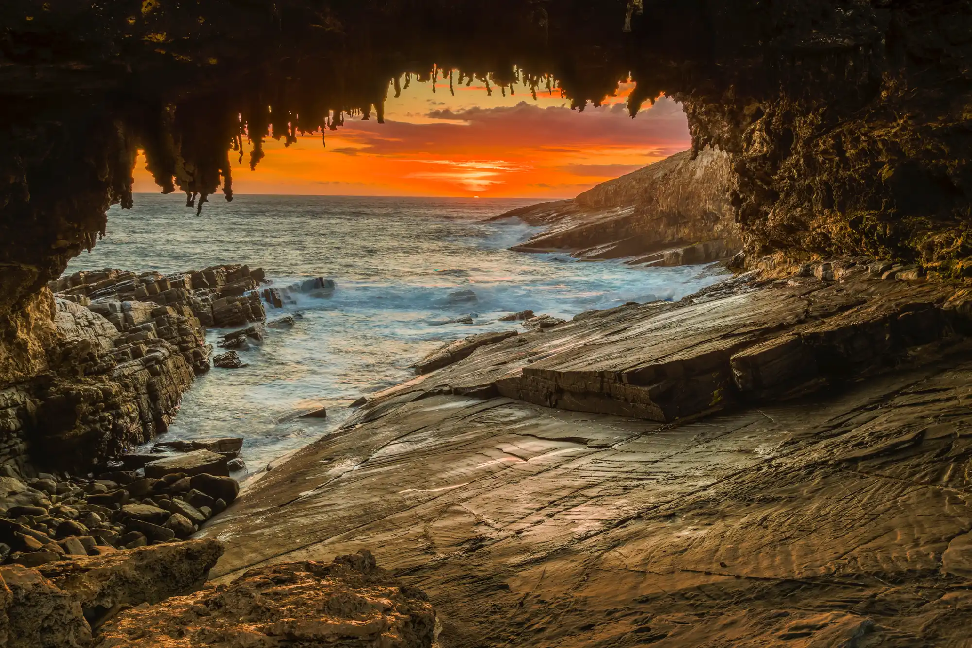 Orange sunset over the Southern Ocean framed by Admirals Arch, showing hanging limestone formations and waves breaking against the rocky Kangaroo Island coastline.