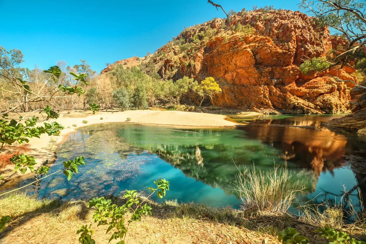 A tranquil, clear green waterhole reflects towering red-orange cliffs under a bright blue sky, with a sandy bank and eucalyptus trees surrounding the water.
