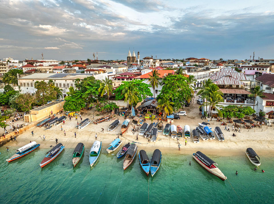 Aerial view of dhow boats moored on the Stone Town waterfront, Zanzibar, with historic buildings and turquoise Indian Ocean waters.