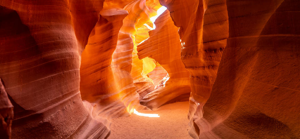 Interior of Lower Antelope Canyon in Arizona with glowing orange sandstone walls, narrow passageways, and sunlight streaming through the canyon opening onto sandy ground.