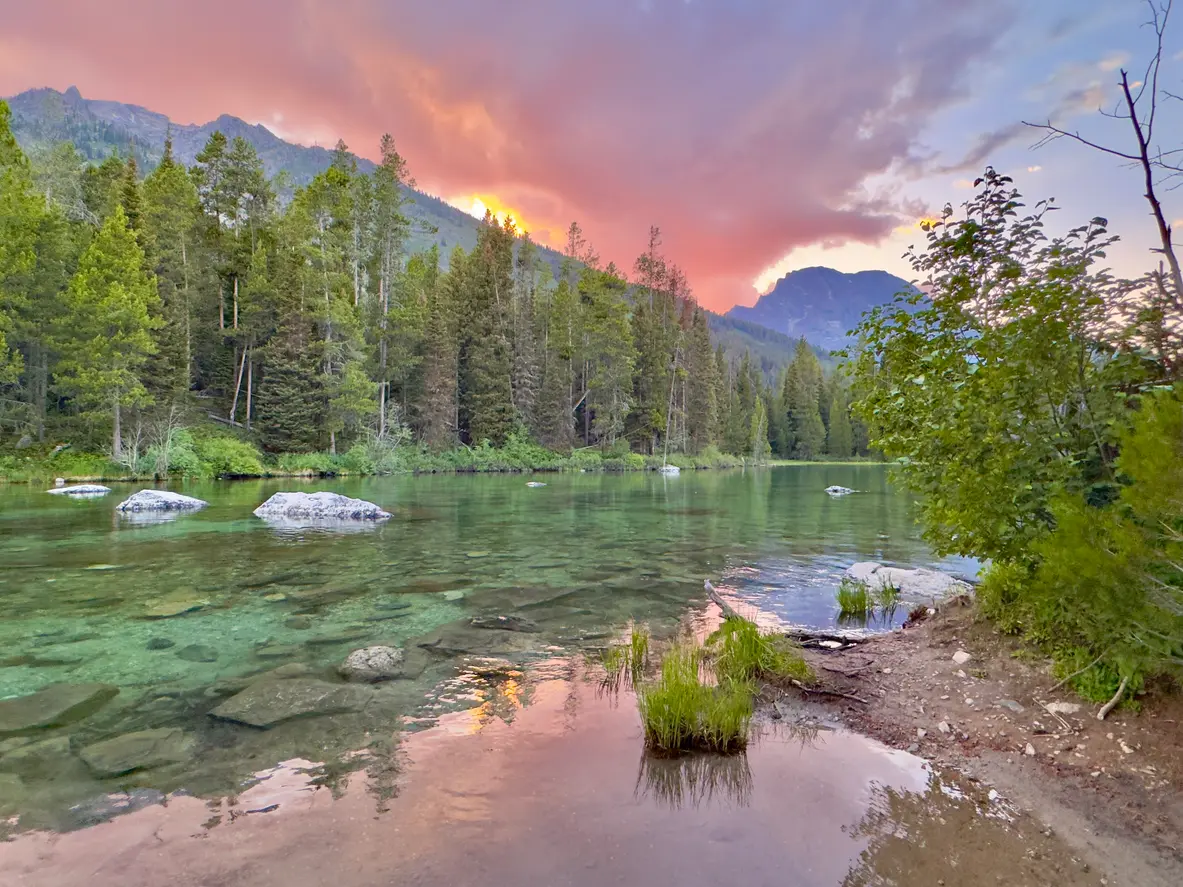 Clear water of String Lake with rocks and forest reflected under pink and purple sunset in Grand Teton National Park, Wyoming, USA.