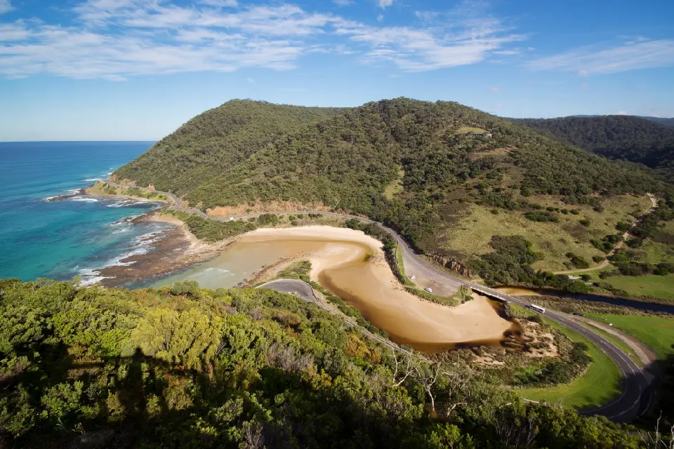 Elevated view of the Great Ocean Road curving around a sandy inlet between the ocean and forested hills near Lorne, Victoria.