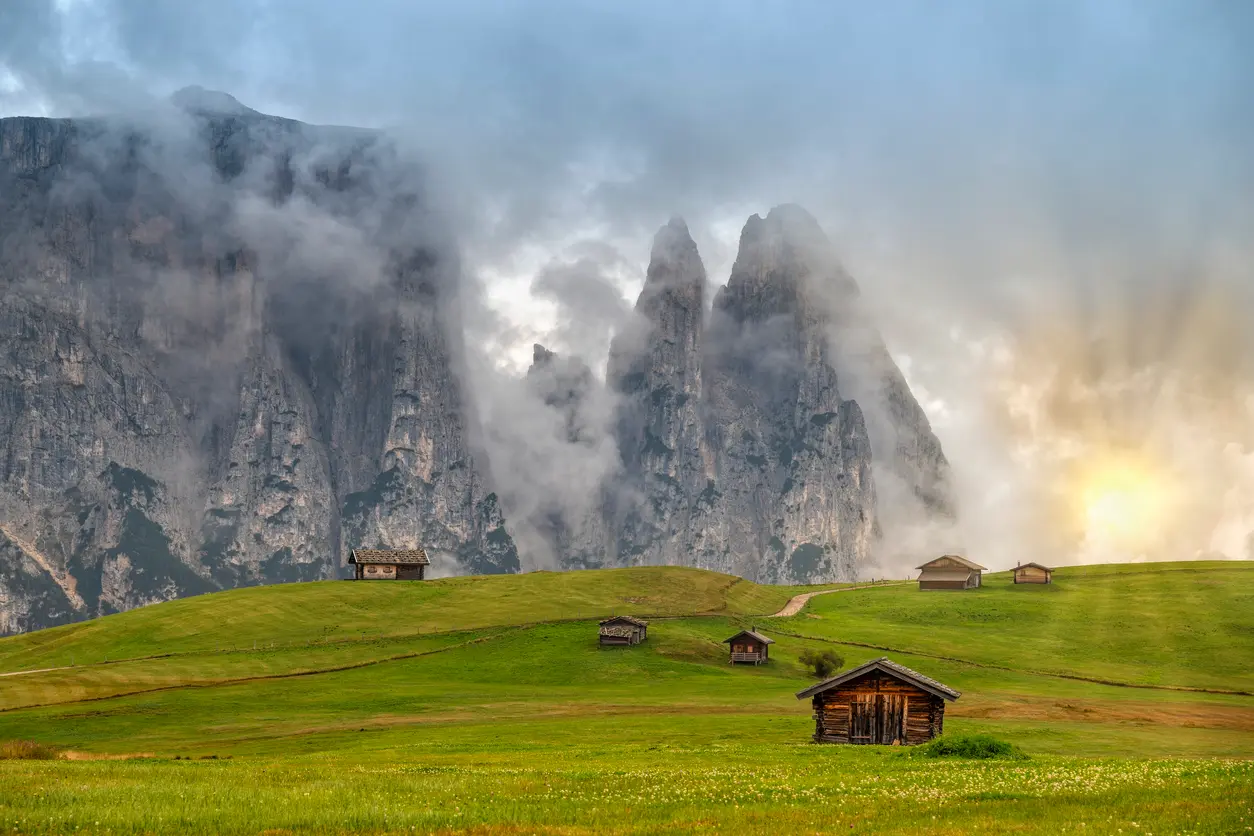 Alpine huts on rolling green meadows of Alpe di Siusi with the Schlern mountains rising behind in the Dolomites, South Tyrol, Italy.