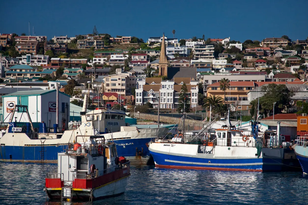 Fishing boats in Mossel Bay Harbour with the coastal town, church spire and hillside neighbourhoods in the background, Western Cape, South Africa.