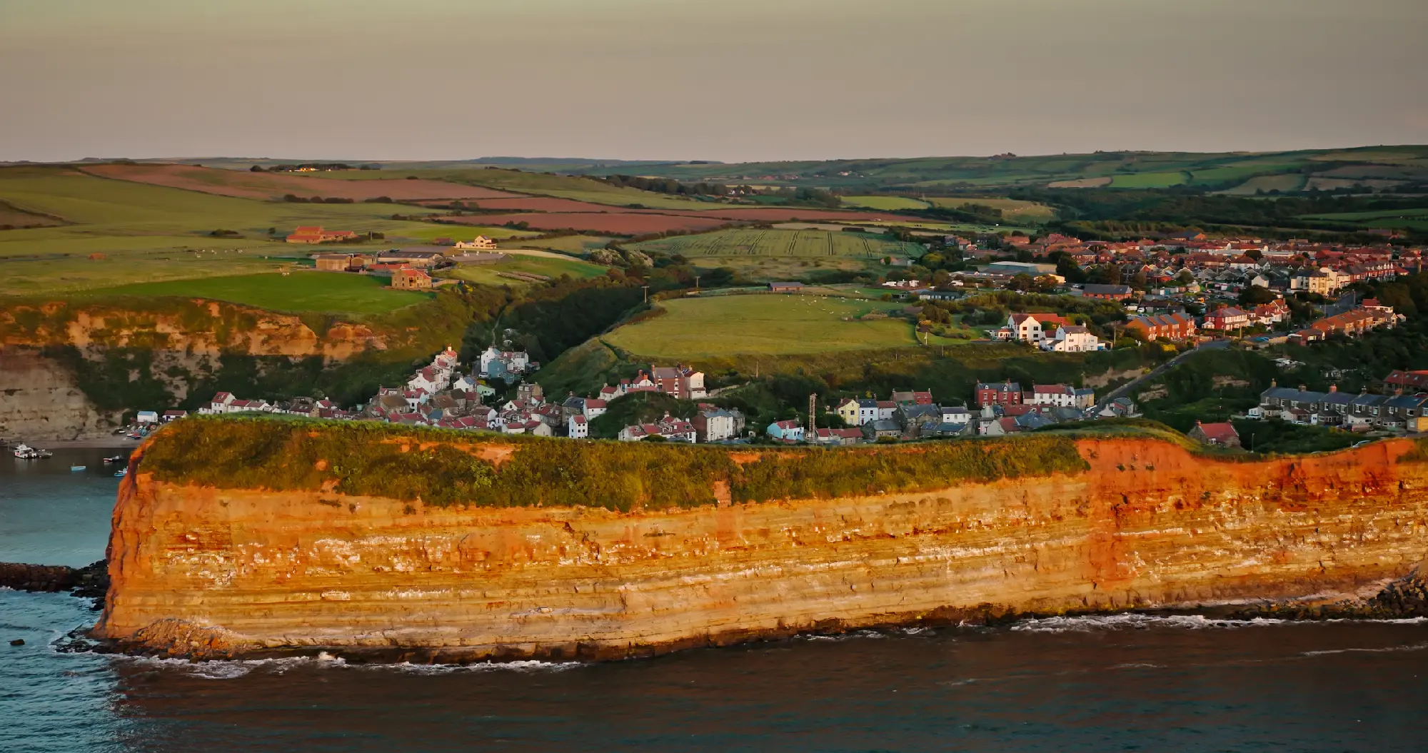 Aerial view of sunlit sandstone cliffs, Staithes fishing village and rolling farmland along the North Yorkshire coast, England.