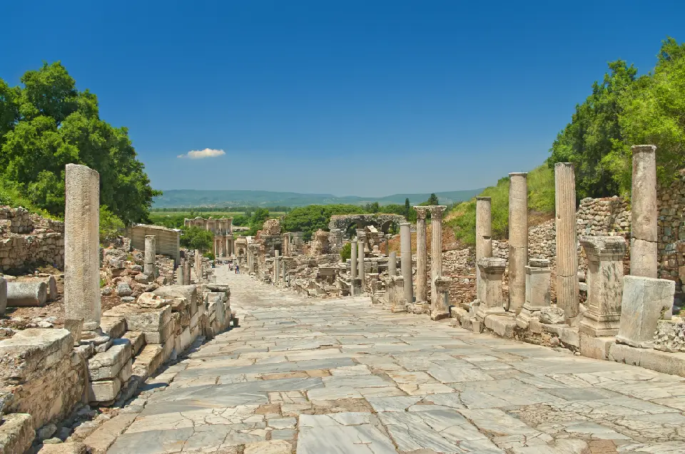 Wide marble street lined with ancient columns and ruins, descending toward the Library of Celsus at Ephesus beneath a clear blue sky.