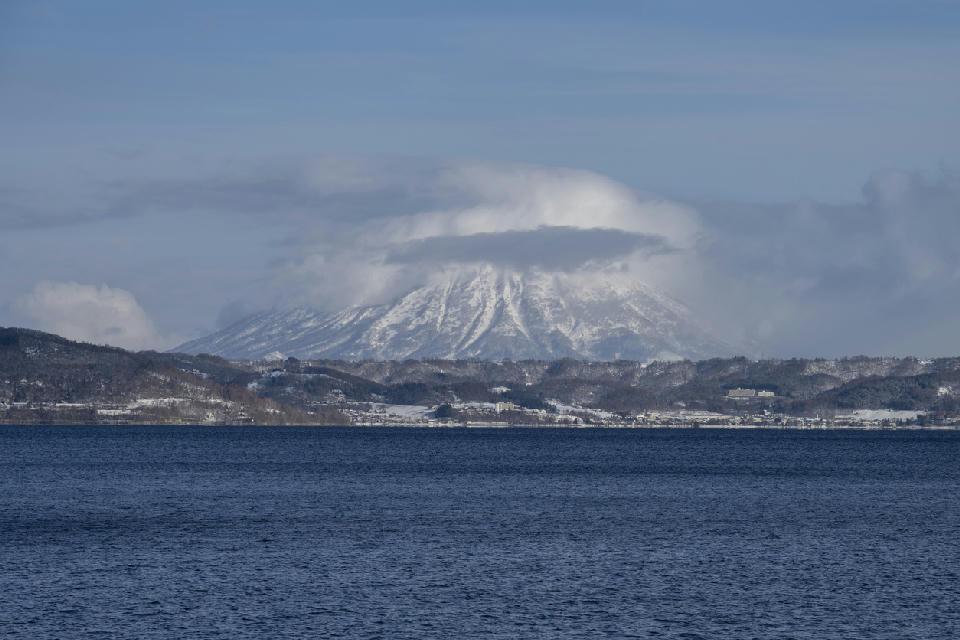 Snow-covered Mount Usu rising beyond Lake Toya with low clouds wrapping the summit, viewed across the lake in Hokkaido, Japan.