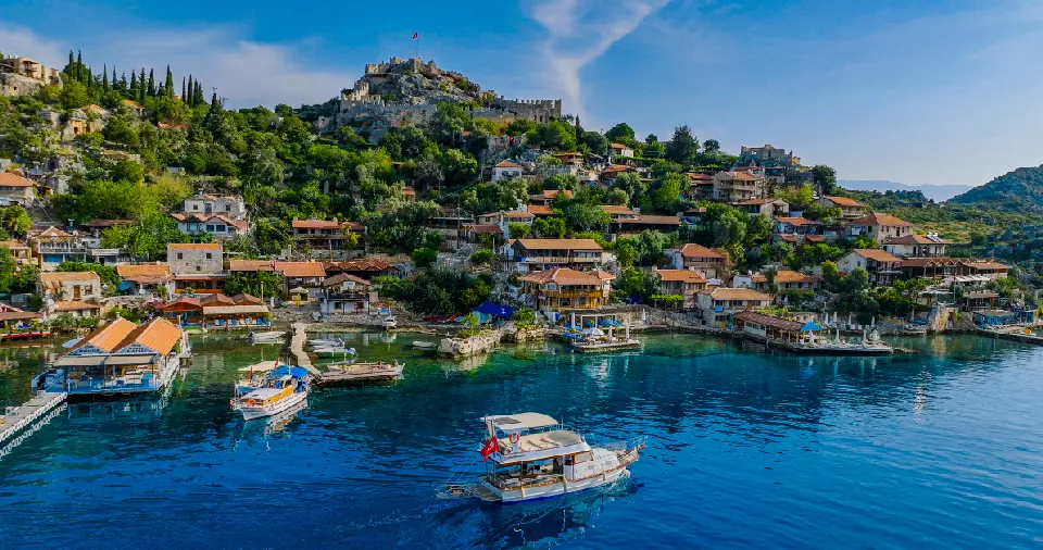 Boats cross the clear blue water in front of Kaleköy, where traditional red-roofed houses climb a green hillside toward the historic stone castle of Simena.