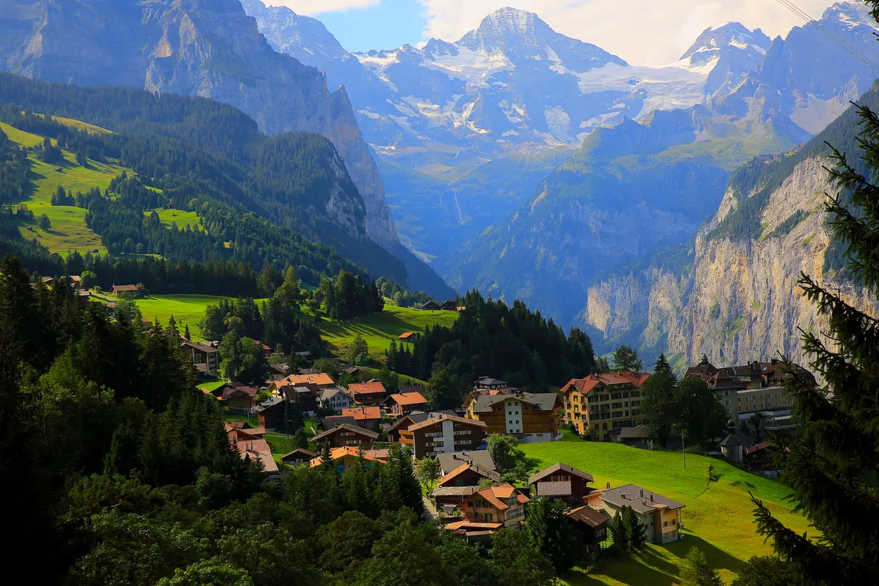 Wengen village on a green hillside overlooking Lauterbrunnen Valley, with steep cliffs and snow-covered alpine peaks in the background, Switzerland.
