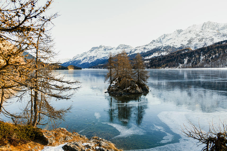 Small island with trees in Lake Sils partially frozen, with snow-covered mountains in the Engadin, Switzerland
