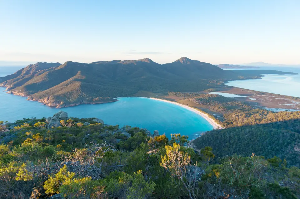 Elevated view over the turquoise waters and sweeping white-sand beach of Wineglass Bay beneath rugged mountains in Freycinet National Park, Tasmania.