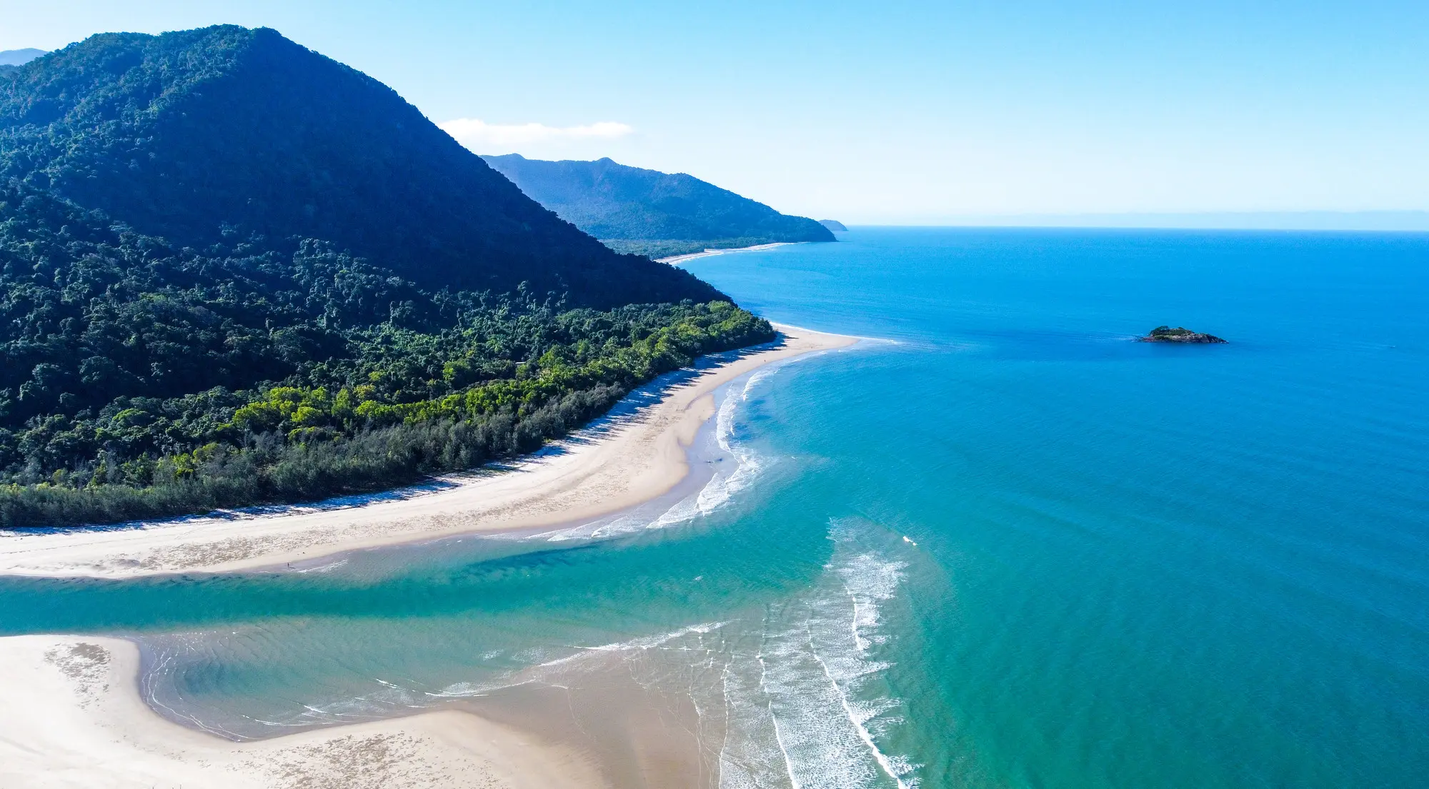 Aerial view of beautiful rainforested mountains sweeping down to a long sandy beach and a bright blue bay of water by the reef.