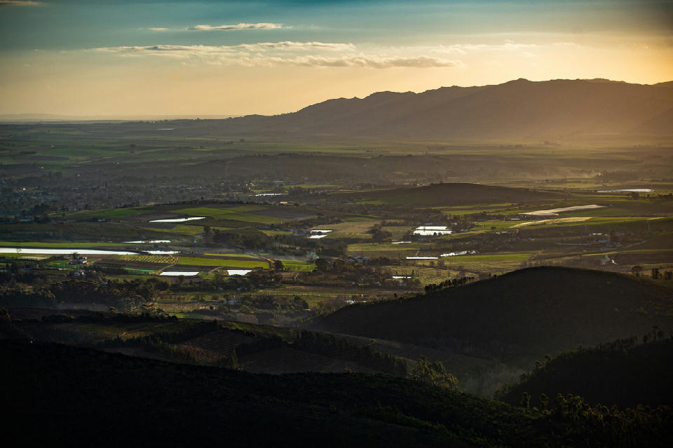 Golden sunset over the Cape Winelands near Wellington, South Africa, showing layered agricultural fields, farm dams, rolling valley terrain, and distant mountain silhouettes.