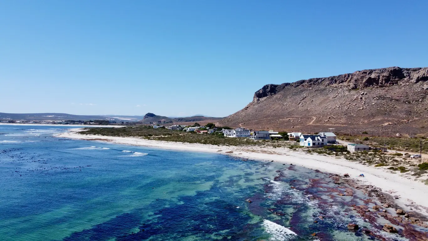 Drone view of Elands Bay on South Africa's West Coast, showing turquoise ocean water, sandy shoreline, coastal houses and the rocky escarpment rising behind the bay.